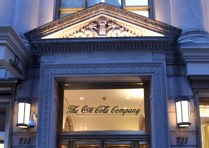 The grand Beaux-Arts entrance of the historic building at 711 Fifth Avenue in New York City, now home to the exclusive Core Club. Ornate stone carvings, classical columns, and decorative pediments frame the illuminated doorway, while the iconic Coca-Cola script remains visible above the entrance as a nod to the building’s storied past. Warm interior lighting contrasts with the blue evening sky, highlighting the landmark architecture along Fifth Avenue.