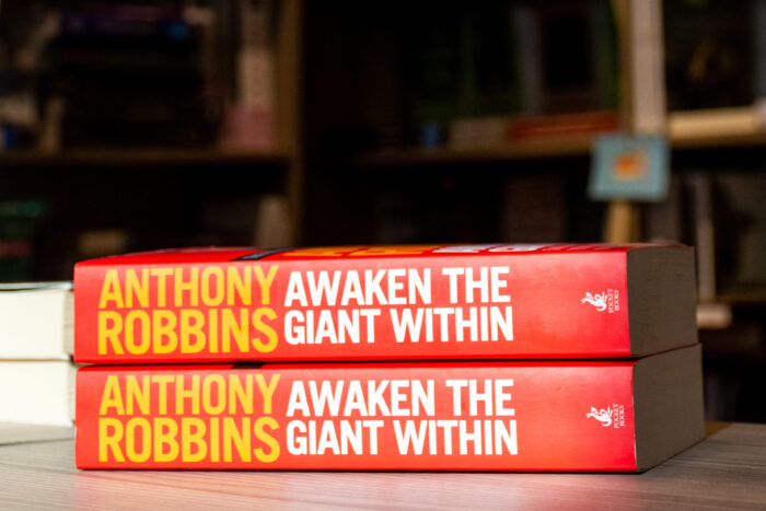Two copies of the book Awaken the Giant Within by Anthony Robbins are stacked on a table, displaying bright red spines with bold yellow and white lettering. The books are positioned in front of softly blurred bookshelves, creating a warm bookstore or library setting. The close-up composition emphasizes the title and self-improvement theme of the bestselling motivational book.