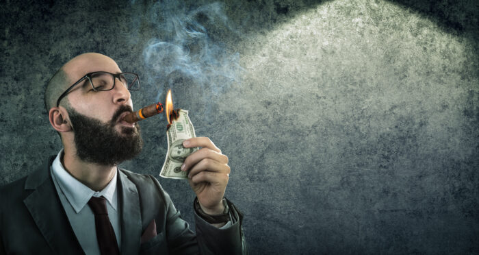 A conceptual image shows a businessman in a suit and glasses lighting a cigar with a burning U.S. hundred-dollar bill. Smoke rises around him against a dark textured background, while a spotlight illuminates the scene. The image symbolizes extravagance, wasteful spending, wealth, excess, or financial recklessness.