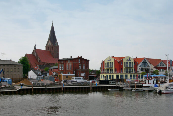 A waterfront view of the harbor in Barth, Germany, with calm water and docked boats in the foreground. Behind the marina, colorful modern buildings and traditional brick structures line the shoreline, while the towering red-brick St. Mary's Church with its tall Gothic spire rises above the town under an overcast sky.