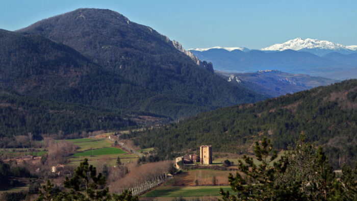A scenic view of the Chateau d'Arques vu vers l'ouest featuring rolling green fields, dense forested hills, and a historic stone tower and church nestled in the valley. In the distance, snow-capped mountains rise above layers of blue ridges beneath a clear sky, creating a dramatic backdrop to the peaceful rural landscape.