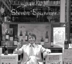 Archival photograph of wine merchant Steven Spurrier seated outside his Paris wine shop, Sélection Steven Spurrier. He raises a glass of wine while surrounded by bottles, reflecting his influential role in organizing the 1976 Judgment of Paris tasting.