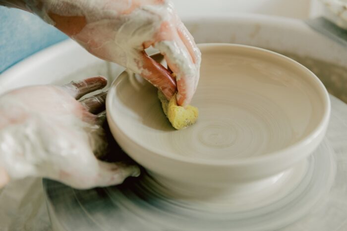 Clay-covered hands refine a handmade ceramic bowl on a spinning pottery wheel. One hand steadies the vessel while the other uses a small sponge to smooth the surface, creating clean lines and an even finish. The close-up composition highlights the precision, patience, and craftsmanship involved in the pottery-making process.