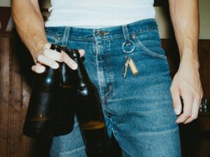 A close-up of a person wearing a white tank top and blue jeans holds three dark glass beer bottles in one hand. A key ring with keys and a lighter hangs from a belt loop, while wood-paneled walls in the background create the atmosphere of a neighborhood dive bar. The candid, flash-lit image has a gritty, nostalgic feel that captures the casual character of nightlife.