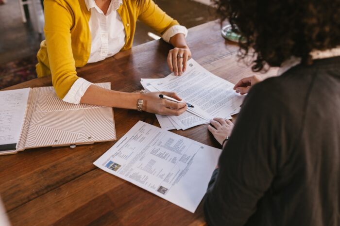 Two people sit across from one another at a wooden table reviewing and signing documents. Papers, a folder, and a pen are spread across the tabletop as they discuss details in a professional office setting.