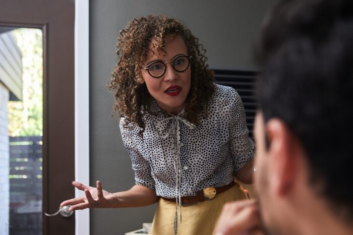 A woman with curly hair and round glasses leans forward during an animated conversation inside a modern home. Dressed in a polka-dot blouse and mustard-colored skirt, she gestures expressively while speaking to a seated person in the foreground. Natural light streams through a nearby glass door, highlighting the intensity and engagement of the exchange.