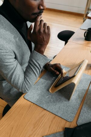 A professional sits at a wooden desk in a modern office, thoughtfully reviewing content on a tablet mounted on a curved wooden stand. Dressed in a gray blazer and black turtleneck, the individual rests a hand against their chin in a contemplative pose. The minimalist workspace features clean lines, natural materials, felt desk pads, and soft natural light, conveying focus, productivity, and modern workplace design.