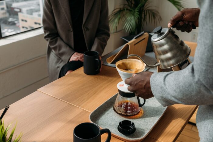 A close-up view of two professionals sharing coffee in a modern office setting. One person pours hot water from a stainless-steel kettle into a pour-over coffee dripper, while another sits across the table holding a black mug. The workspace features light wood furnishings, indoor plants, large windows with city views, and a relaxed atmosphere that emphasizes connection, hospitality, and workplace culture.