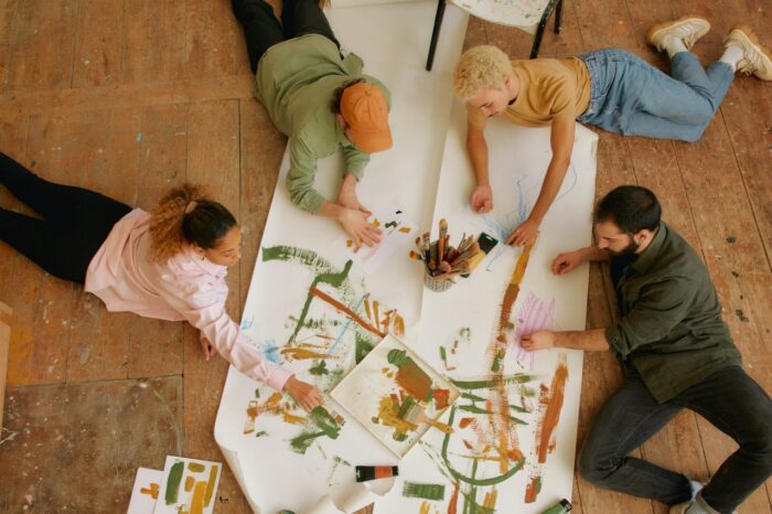 An overhead view shows four people lying and kneeling around a large sheet of paper spread across a wooden floor, collaborating on a colorful abstract painting. Paintbrushes, trays of paint, and scattered sketches surround the group as they create bold strokes in green, orange, blue, and pink. The scene captures a relaxed, creative atmosphere centered on teamwork, artistic expression, and shared inspiration.