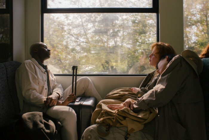 Three travelers sit quietly across from one another on a train, bathed in warm afternoon light streaming through a large window. One person rests against a suitcase while gazing upward, while two companions lean together in a moment of calm reflection. Trees blur softly outside, creating a peaceful atmosphere that evokes slow travel, companionship, and contemplation.