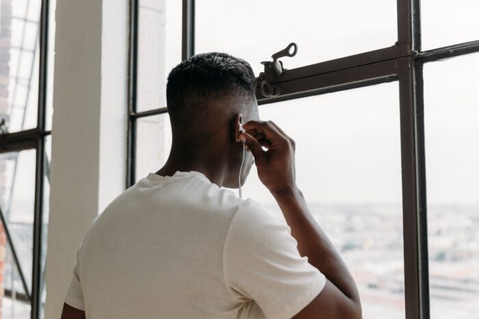 A person stands beside a large industrial-style window, adjusting an earbud while looking out over a blurred city skyline. Natural light fills the modern loft-like space, highlighting the clean lines of the window frame and the minimalist setting. The image conveys a quiet moment of reflection, focus and connection in a contemporary remote-work environment.