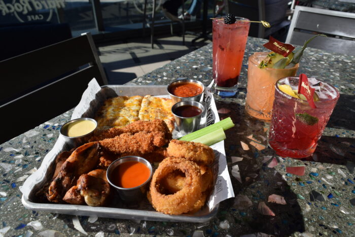 A platter of appetizers and handcrafted cocktails sits on a terrazzo-topped patio table at Hard Rock Cafe. The appetizer sampler includes chicken wings, mozzarella sticks, onion rings, celery sticks, toasted bread and a cheese-covered flatbread, accompanied by an assortment of dipping sauces. Four colorful cocktails garnished with fresh fruit and branded cocktail picks are arranged beside the tray. Sunlight casts dramatic shadows across the table, creating a vibrant outdoor dining scene.