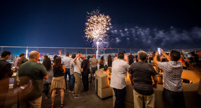 A crowd gathers on a rooftop terrace overlooking the Las Vegas Strip as bright fireworks explode in the night sky. Guests watch and record the display on their phones while the illuminated skyline stretches across the horizon, creating a festive atmosphere.