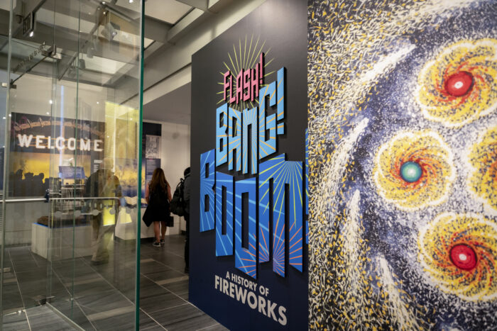 Interior of a museum entrance featuring a large, colorful exhibition sign reading “Flash! Bang! Boom! A History of Fireworks.” The bold blue and pink lettering stands beside a vibrant abstract artwork depicting bursts of fireworks in yellow, red, and blue. Through the glass entryway, visitors walk toward a warmly lit gallery with a glowing “Welcome” sign visible in the background.