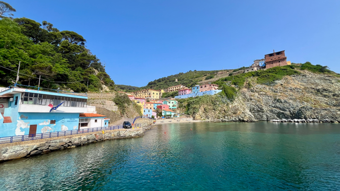The small harbor of Gorgona Island features colorful pastel buildings nestled into a rocky hillside above turquoise water under a bright blue sky.