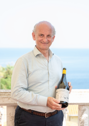 Lamberto Frescobaldi stands on a seaside terrace holding a bottle of Gorgona wine, with the Mediterranean Sea softly blurred in the background.
