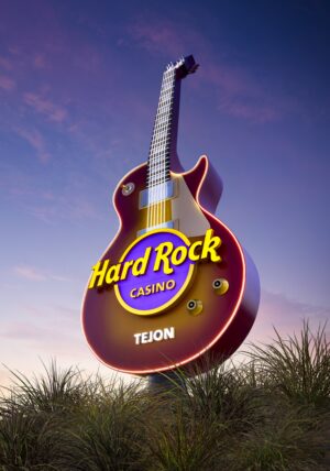 A towering guitar-shaped monument stands against a twilight sky at the entrance to Hard Rock Hotel & Casino Tejon. The oversized electric guitar, finished in deep red with illuminated edges, features the iconic Hard Rock logo at its center and the word “Tejon” near the base. Ornamental grasses surround the monument, while soft purple and blue hues color the evening sky, creating a dramatic and visually striking landmark.
