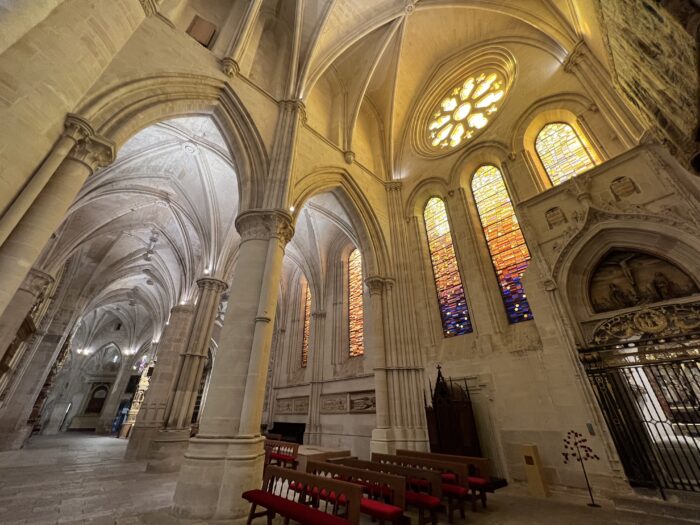 Interior of a Gothic cathedral featuring soaring ribbed vaults, massive stone columns, and pointed arches illuminated by warm light. Tall stained-glass windows in shades of gold, amber, and deep blue cast colorful reflections across the pale stone walls, while rows of wooden pews with red cushions sit beneath the vaulted ceiling.