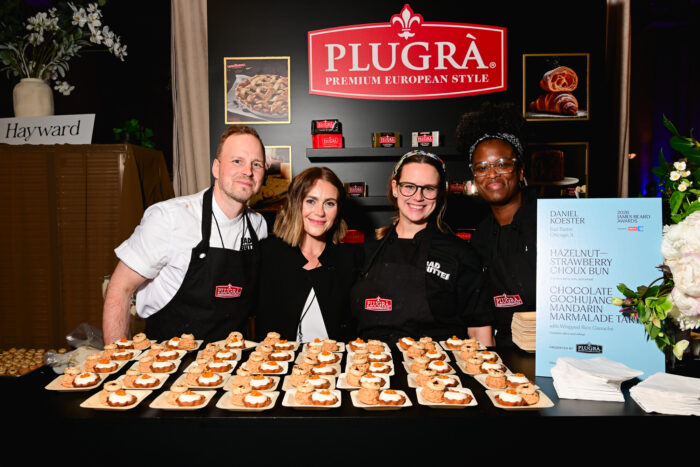 A pastry team from Bad Butter in Chicago poses behind a display of miniature desserts during the 2026 James Beard Awards reception. Trays of hazelnut-strawberry choux buns and chocolate gochujang mandarin tarts are arranged in front of a Plugrà Premium European Style Butter display, celebrating the craftsmanship behind the evening's culinary offerings.