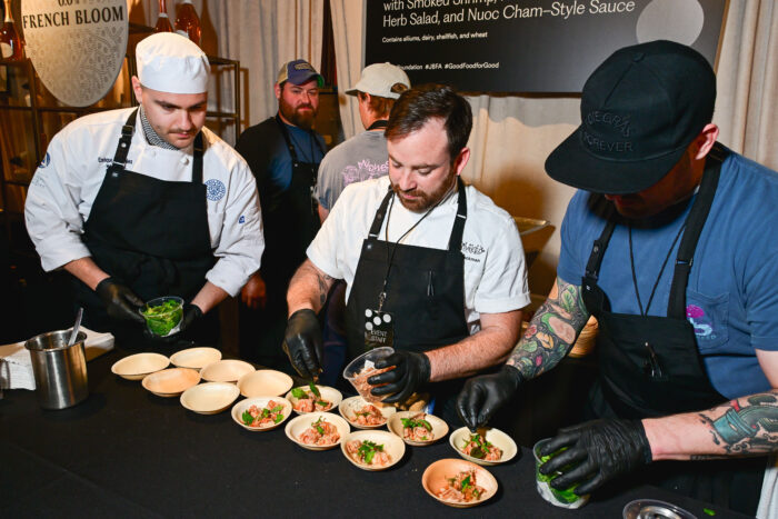 Chefs work together behind the scenes plating smoked shrimp appetizers during the 2026 James Beard Awards reception. Wearing chef coats, aprons, and gloves, the culinary team carefully garnishes each dish before service, highlighting the precision and collaboration that define the annual celebration of American food and hospitality.