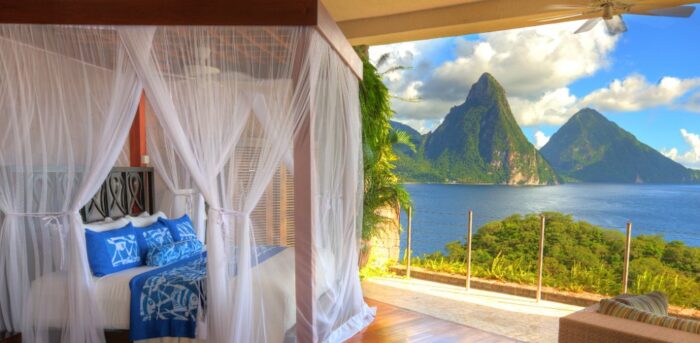 A luxurious open-air resort suite overlooks a brilliant blue Caribbean bay framed by two dramatic volcanic peaks. A canopy bed draped in sheer white netting sits on a polished wood floor, accented with blue-and-white linens. Sunlight streams into the room, highlighting lush tropical greenery and panoramic views of the sea and mountains beyond.