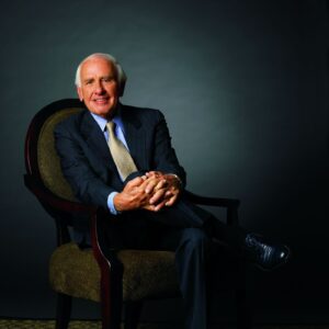 A professionally lit studio portrait of Jim Rohn seated in an upholstered chair against a dark blue-gray background. Dressed in a navy suit, light blue shirt, and patterned tie, he smiles warmly at the camera with his hands clasped in his lap. The formal composition and subdued backdrop create a polished, executive-style portrait.