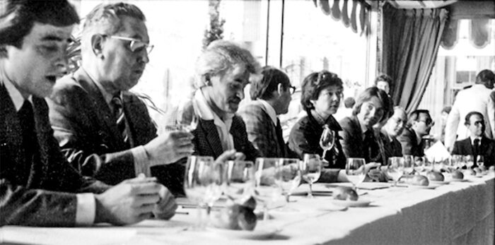 Historic black-and-white photograph of French wine judges seated at a long tasting table during the 1976 Judgment of Paris. Glasses, score sheets, and bottles are arranged before the panel as they evaluate wines in a blind tasting.