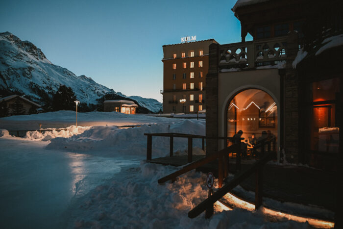 The Kulm Country Club in St. Moritz, Switzerland, glows warmly against a snowy alpine landscape at dusk. Guests gather inside the illuminated clubhouse while snow-covered mountains rise in the background. The scene captures the cozy elegance of a winter evening in the Swiss Alps, with soft lights reflecting across the ice and fresh snow surrounding the historic resort.