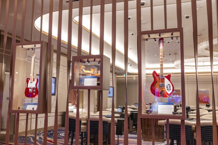 Glass display cases showcasing music memorabilia stand within the casino floor of Hard Rock Hotel & Casino. Featured items include signed guitars and personal artifacts from renowned musicians, illuminated by spotlights inside sleek display boxes mounted among tall decorative metal slats. Plush gaming tables and upholstered chairs fill the background, while warm lighting and contemporary finishes create an upscale atmosphere that blends entertainment, gaming and rock-and-roll history.