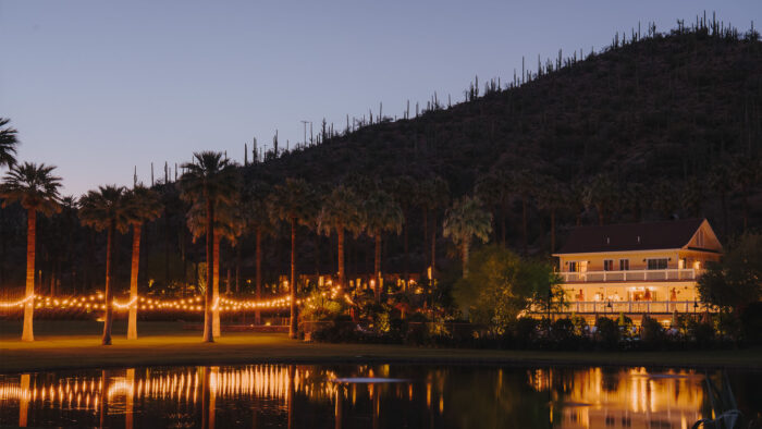 A warmly lit lodge glows against the desert landscape at dusk, surrounded by towering palm trees and strings of twinkling lights. The illuminated building overlooks a still pond that reflects the golden lights and evening sky. In the background, a hillside dotted with cacti rises into the fading twilight, creating a serene oasis atmosphere.