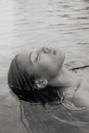 A black-and-white photograph captures a person floating serenely on calm water with eyes closed and face turned toward the sky. Gentle ripples surround them, conveying stillness, surrender, and quiet contemplation.