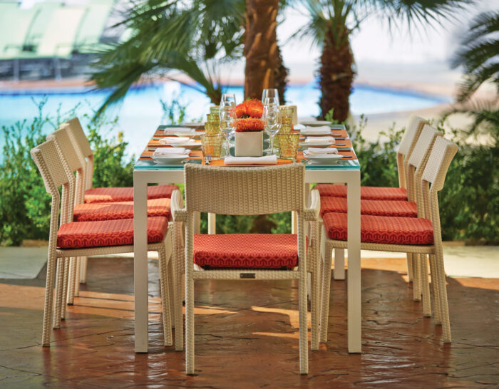An elegantly set outdoor dining table overlooks a resort pool framed by palm trees. Eight wicker chairs with vibrant orange cushions surround a glass-topped table arranged with white place settings, amber glassware and bright floral centerpieces, creating a warm, upscale atmosphere for al fresco dining.