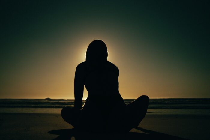 A silhouetted person sits cross-legged on a quiet beach facing the ocean at sunset, with the sun glowing directly behind them and creating a soft halo around their figure. Calm waves, a darkened shoreline, and a clear evening sky evoke a peaceful, contemplative atmosphere.