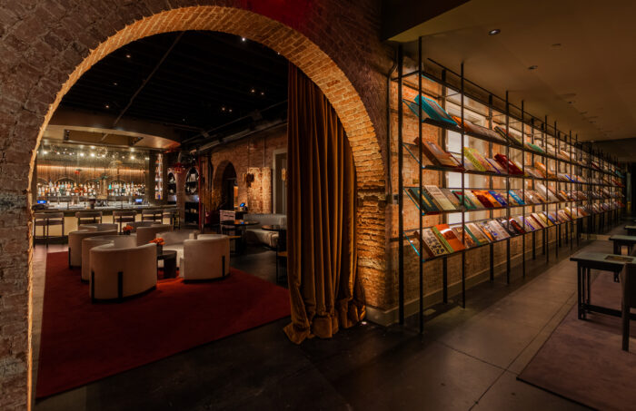 A warmly lit, members-only lounge features exposed brick archways, plush cream-colored seating, and a sophisticated cocktail bar in the background. Along one wall, a striking floor-to-ceiling display showcases hundreds of colorful books and magazines on black metal shelving. Rich textures, dramatic lighting, and intimate seating areas create an atmosphere that feels equal parts private library, luxury hotel lounge, and social club.
