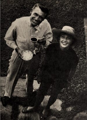 Actor Cary Grant and actress Betsy Drake smile for a candid black-and-white photograph outdoors, with Grant holding a teacup and saucer while Drake wears a wide-brimmed hat beside a garden hedge.