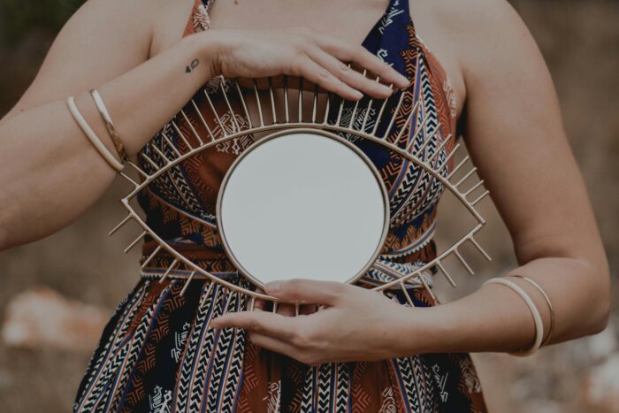 A person wearing a patterned sleeveless dress holds a decorative eye-shaped mirror with a round reflective center and radiating metal accents. Their hands rest gently on the frame, with minimalist bracelets and a small wrist tattoo visible against a softly blurred outdoor background.