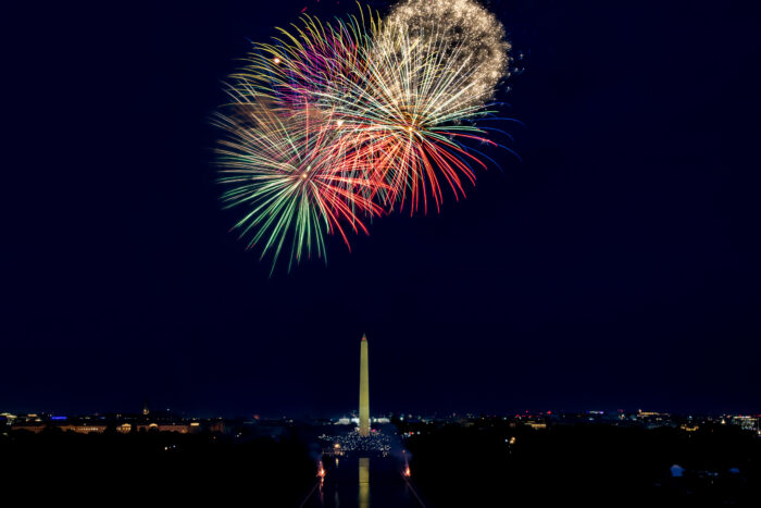 Brilliant red, green, gold, and white fireworks burst above the Washington Monument during an Independence Day celebration in Washington, D.C. The colorful display fills the night sky while the illuminated monument stands centered below, framed by the National Mall and the city skyline.