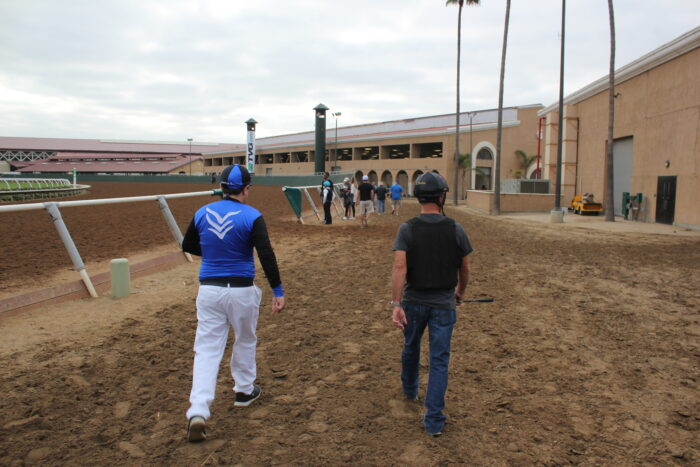 Corey Levitan, dressed in blue-and-white jockey silks, walks alongside trainer Gary Stevens on the dirt track at a horse racing facility. Several people stand ahead near the rail as they make their way toward the training area, with stable buildings and grandstand structures visible in the background.