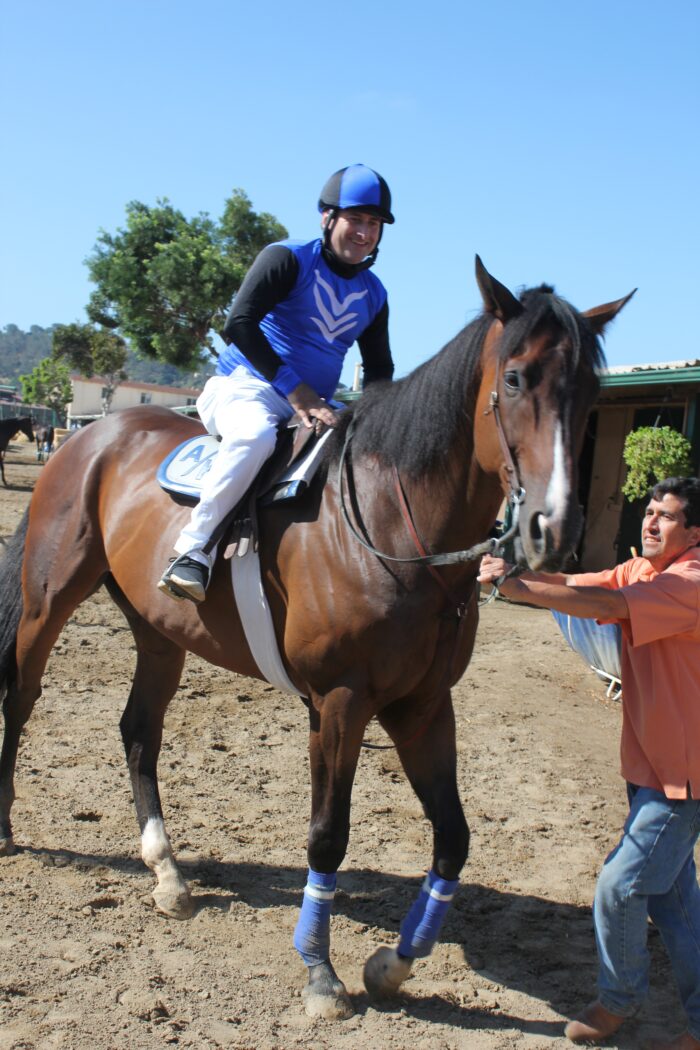Smiling from the saddle, Corey Levitan rides a bay racehorse while a handler in an orange shirt steadies the horse by the reins. The scene takes place in a stable area with other horses and barns visible in the background.