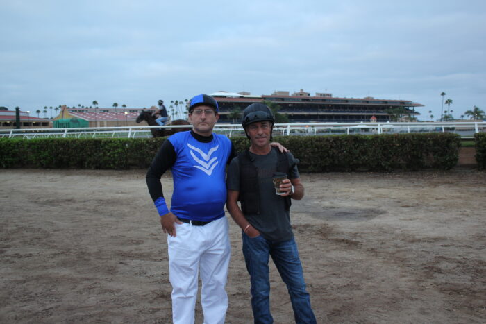 Corey Levitan poses with a horse racing trainer Gary Stevens near the track at dusk. A racehorse and rider pass in the background, with the grandstand and racetrack illuminated under a cloudy evening sky.