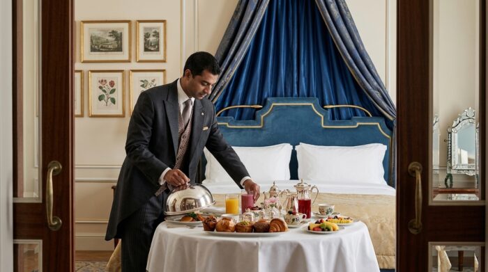 AI-generated image of a hotel butler in formal attire prepares an elegant in-room breakfast beside a plush blue canopied bed. The round table is set with fresh pastries, fruit, tea service and juices, illustrating the personalized hospitality and attentive service associated with luxury accommodations.
