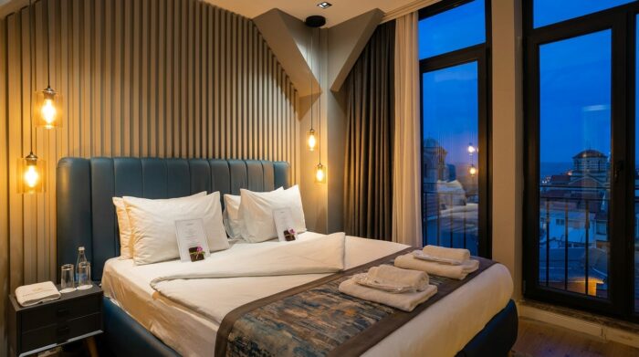 AI-assisted image of a luxury hotel room prepared for evening turndown service features a neatly folded duvet, plush towels placed on the bed and chocolates resting atop personalized cards. Warm pendant lighting and floor-to-ceiling windows overlooking a city skyline at dusk create an inviting and tranquil atmosphere.