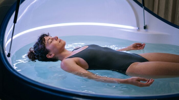 An AI-generated illustration shows a person floating peacefully in a sensory deprivation tank with eyes closed and arms relaxed at their sides. Soft blue lighting reflects across the still water, creating a calm, restorative atmosphere associated with float therapy and deep relaxation.