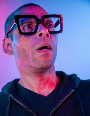 Close-up portrait of Ernest Hemmings wearing oversized black square glasses, illuminated by vibrant pink and blue lighting. He looks upward with a surprised expression, his mouth slightly open, against a softly blurred neon-colored background.