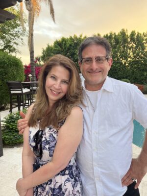 Jo Ann and Corey Levitan smile for a portrait beside a backyard pool at sunset, with lush greenery and palm trees in the background, reflecting the warmth and companionship of their long-term marriage.