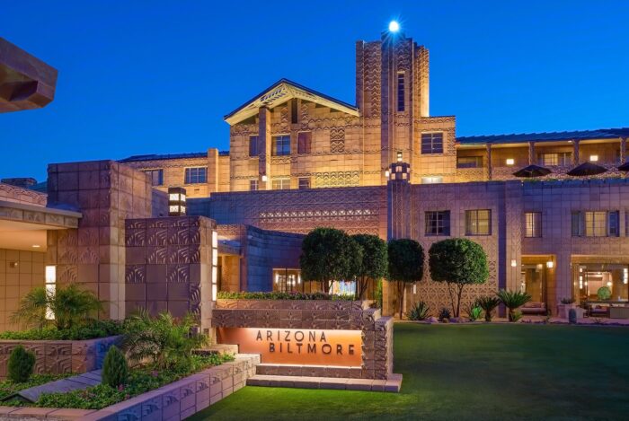 Arizona Biltmore at dusk, showcasing its iconic Frank Lloyd Wright-inspired architecture, illuminated stone façade, manicured gardens, and dramatic Art Deco tower against a deep blue evening sky.