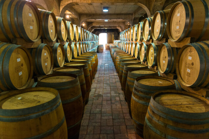 Rows of oak wine barrels line both sides of a brick-floored cellar, stacked two high beneath concrete arches. Warm lighting leads the eye toward a bright doorway at the far end, highlighting the symmetry and depth of the aging room.