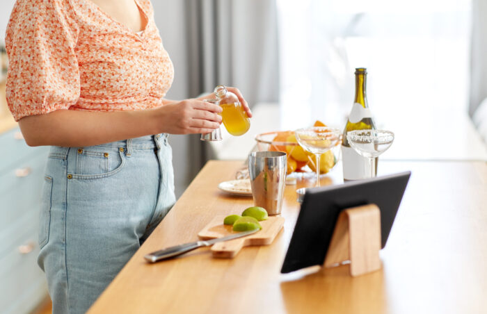 drinks and people concept - close up of woman with tablet pc computer pouring liquor or syrup into jigger and making cocktail at home kitchen