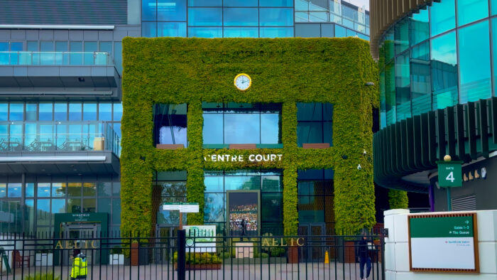 Exterior view of Wimbledon’s Centre Court entrance, featuring a striking facade covered in lush green ivy. The iconic clock sits above the entrance, with modern glass buildings framing the scene and directional signs for the tournament grounds visible in the foreground.