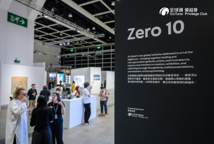 Interior view of the Zero 10 exhibition at Art Basel Hong Kong. A large black sign in the foreground introduces the exhibition, while visitors explore contemporary artworks displayed in bright white gallery booths beneath an industrial-style ceiling with geometric lighting. Conversations among attendees create the atmosphere of a busy international art fair.